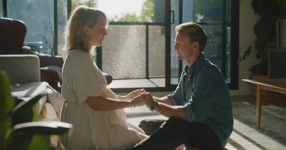 A man and woman sit on the floor at home and hold hands and close their eyes as they meditate