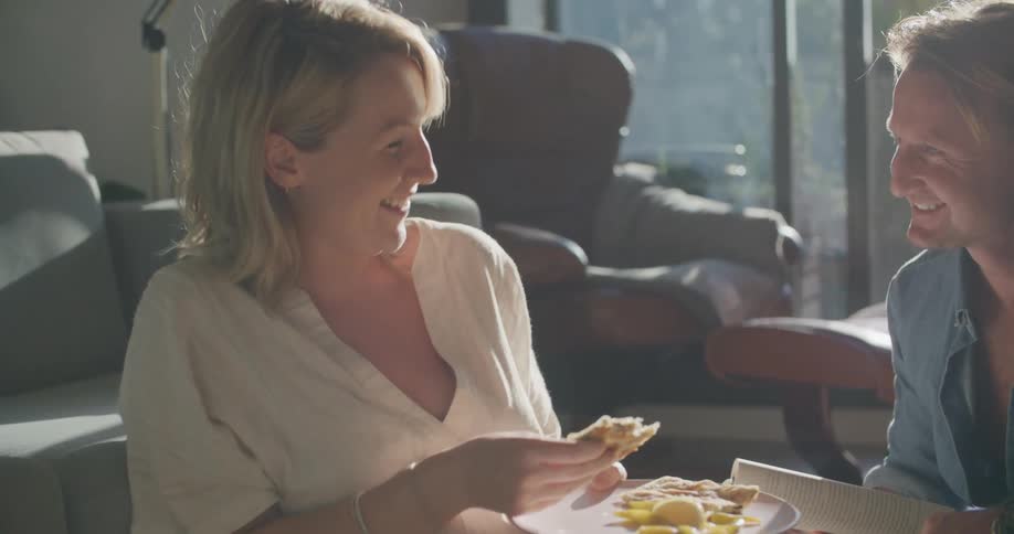 A man and woman sit on the floor in a living room while chatting as the woman eats food from a plate
