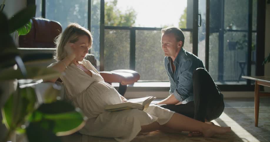 A man sits on the floor sipping coffee and smiling as he looks at his pregnant wife