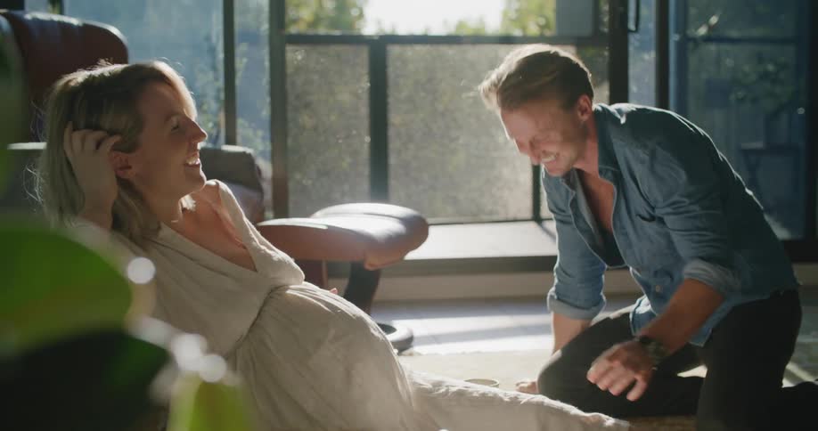 A woman in a room sitting on the floor as a man touches and kisses her on her pregnant stomach with a couch in the background