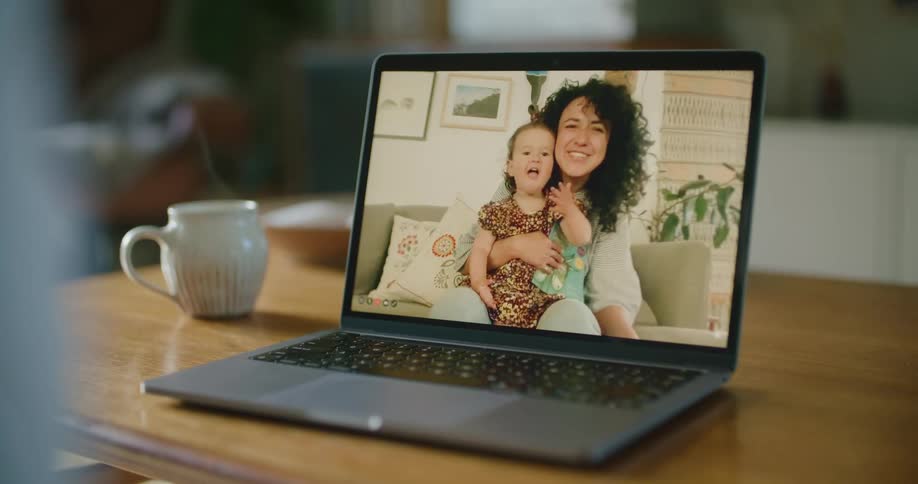 Person makes a video call on laptop with woman holding a toddler girl sitting on the sofa in the living room
