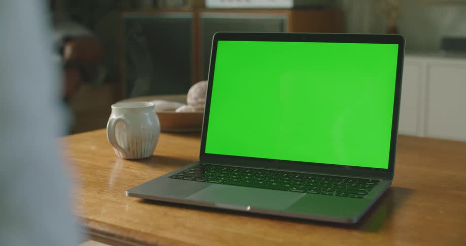 A laptop in a room on a table next to a mug with a hot smoking drink