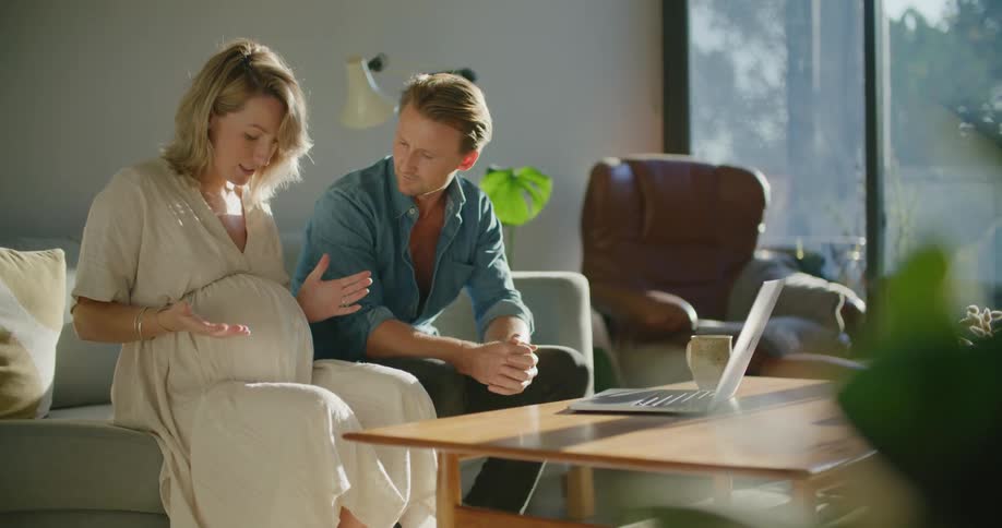A man puts his hand on his wifes pregnant belly as they sit side by side on their sofa with the laptop open on the coffee table