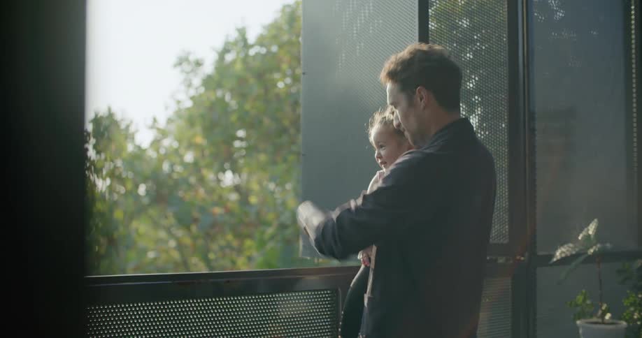 A man holds his toddler daughter out on a balcony and they look at the scenery