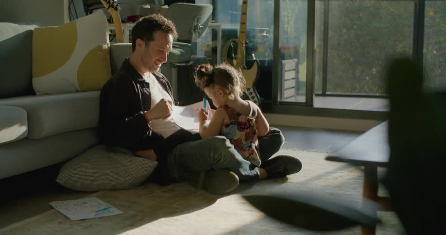 Father sits with daughter on floor in living room near guitars as she draws