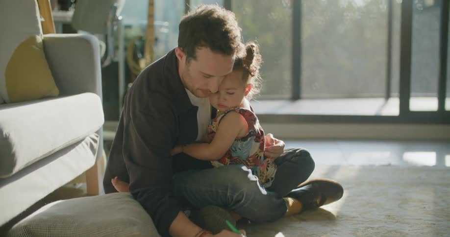Father holding his daughter in his arms draws on paper in the floor of the living room at sunset