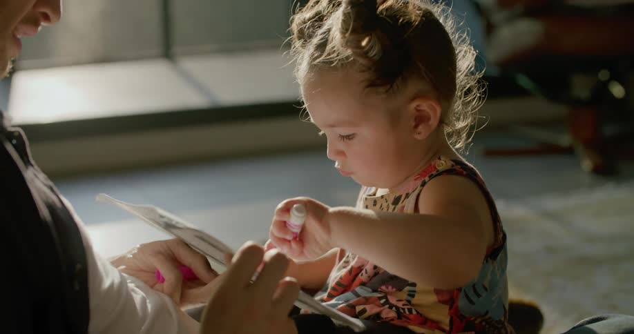 A toddler girl drawing with a marker while sitting on a living room floor with her dad