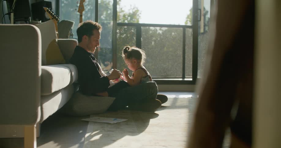A father sitting with his young daughter on a living room floor on a sunny day