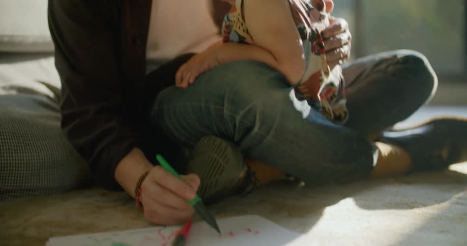 A dad sitting on the floor holding his daughter in his lap as he draws using markers