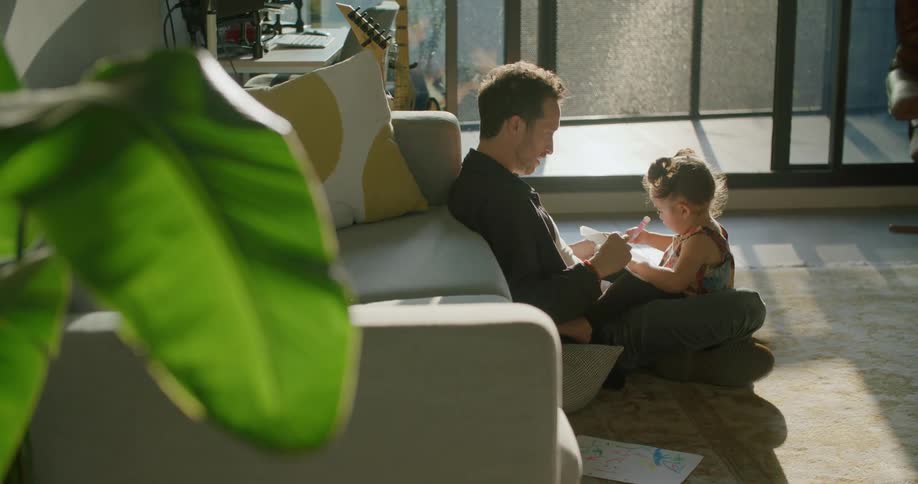 A little girl sits on her dads lap on the floor coloring