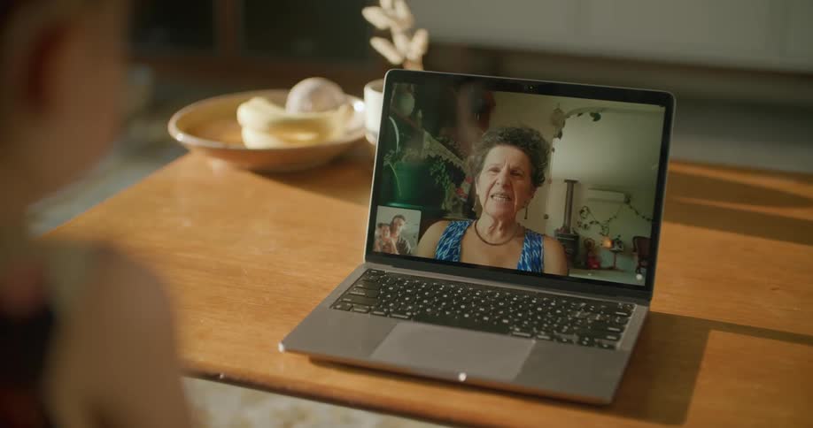 A child sitting by a table in a room video chatting on a laptop with a wall in the background