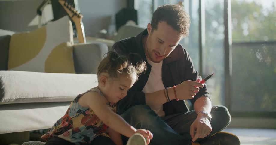 A man sits on the floor and watches as his daughter draws on a paper