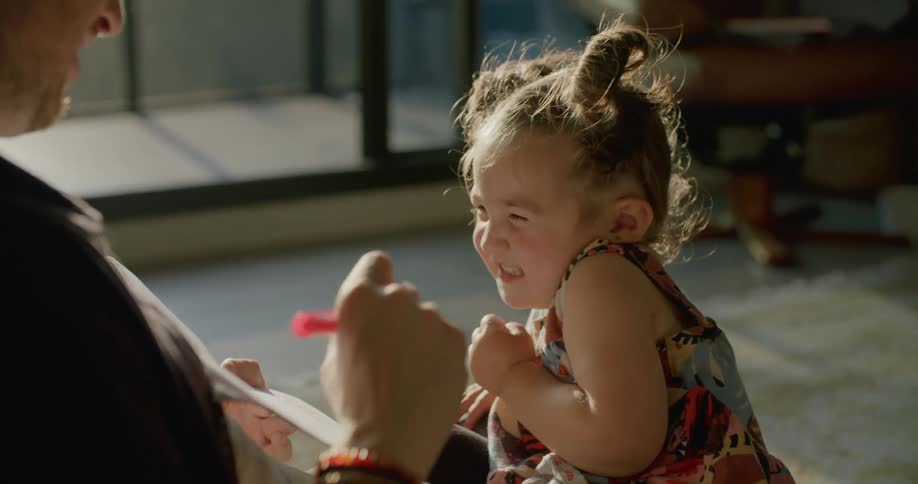 A little girl laughs as she sits on the floor with her dad and he draws on a paper and then puts the felt on her nose