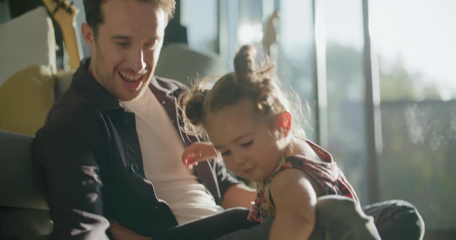 Father holding his daughter in his arms while she draws on paper in the floor of the living room at sunset