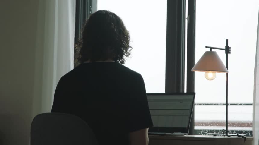 A person in a room sitting on a chair at a desk with a laptop and lamp
