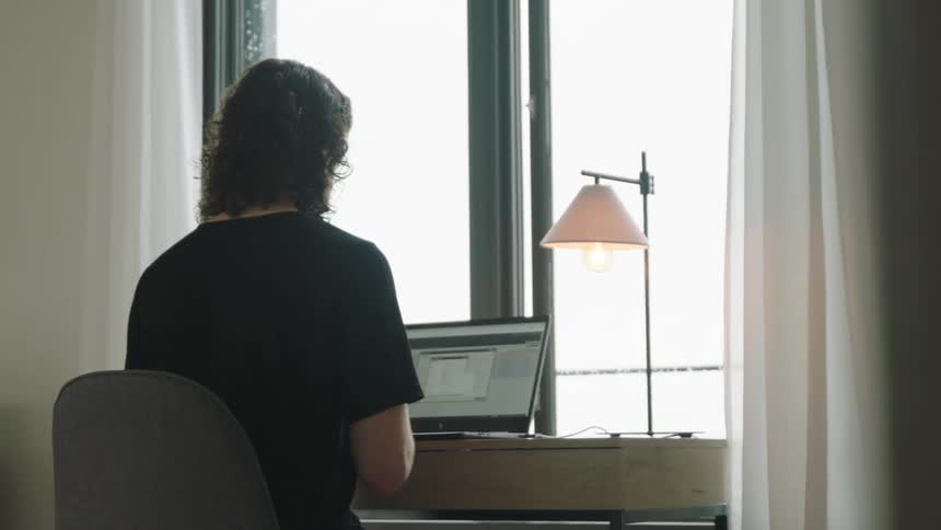 A person sits at a desk in front of a window working on a laptop