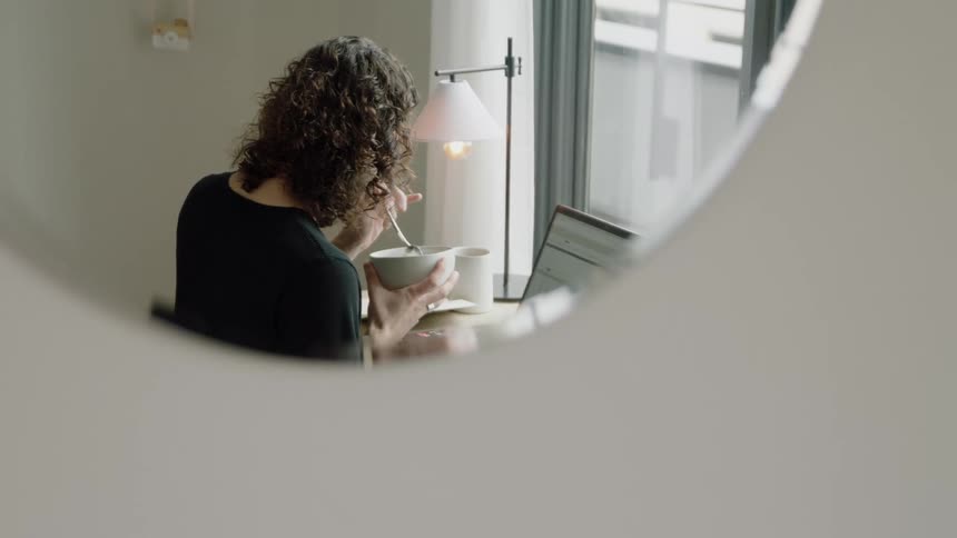 A mirror reflects a woman eating from a bowl with a spoon while sitting before a laptop at a desk during the day