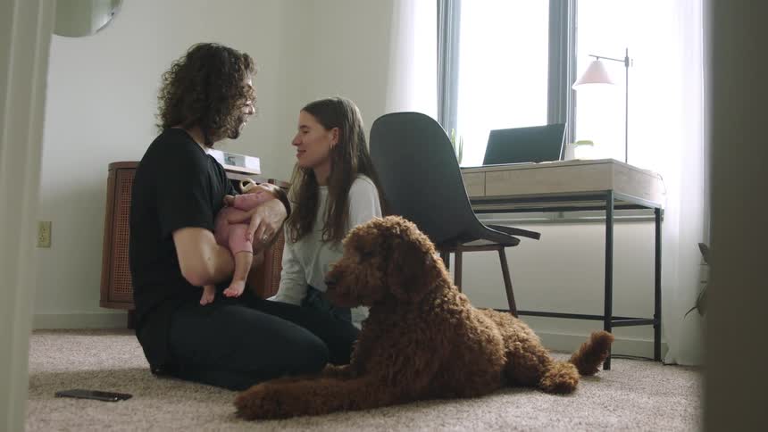 A man and woman sitting on the floor and kissing as the man holds a baby with a dog laying next to them