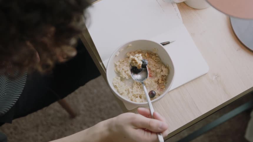A person eats oatmeal with a spoon from bowl lying atop a notebook on a table indoors during the day