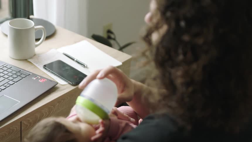 A man sitting on a chair holding a baby and feeding her a bottle next to a desk with a laptop and smartphone