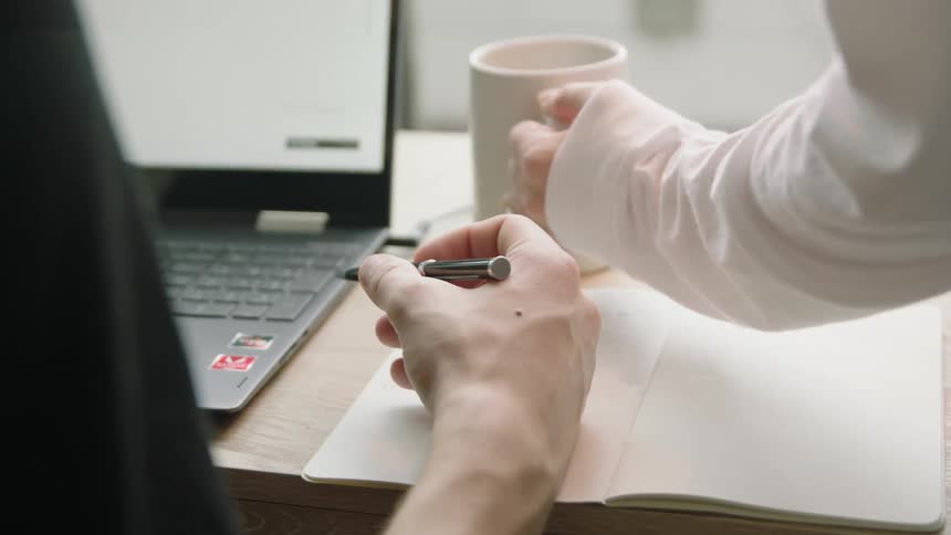 A woman putting a cup of coffee on a mans desk while he writes in a notebook