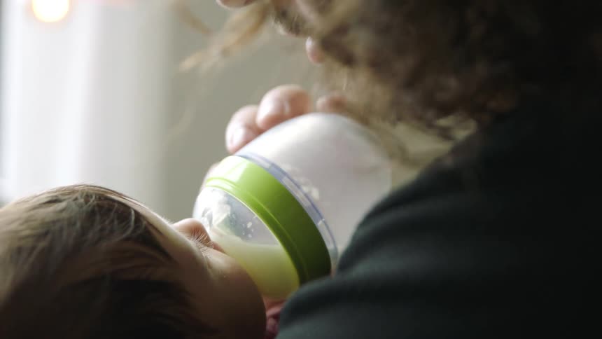 A man kisses the forehead of a baby girl while feeding her with a bottle