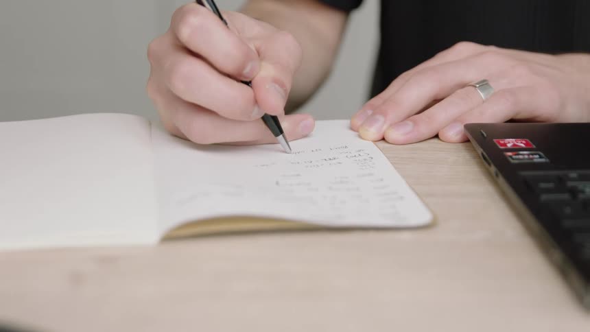 A man sits in front of his laptop and writes in a notebook