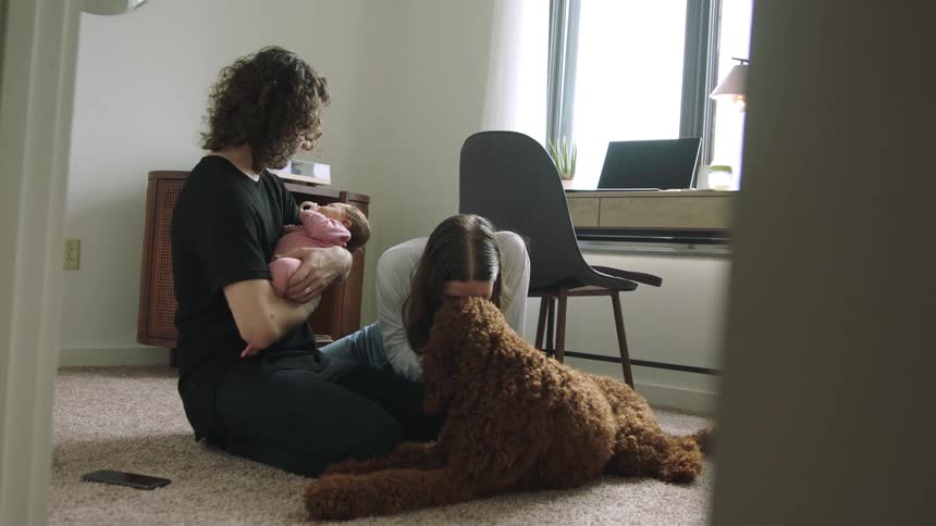 A woman pets and kisses a dog before turning to kiss a baby being held by a man sitting next to her on the floor of a room in a house during the day