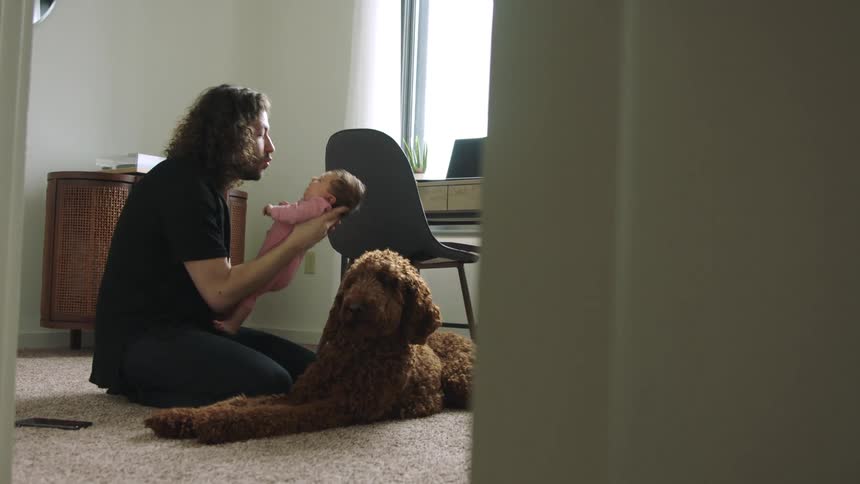 Father sits on floor near dog holding and kissingbaby daughter