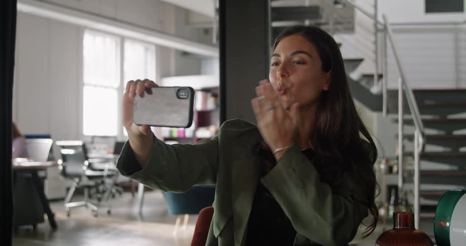 A woman waves and blows a kiss during a video call while sitting at work