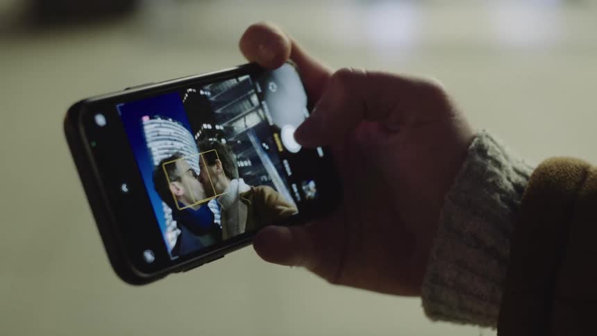 A thumb taps a camera phone button to snap several selfies and the screen shows two men smiling and kissing