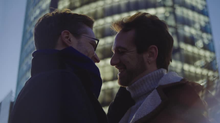 Two men kiss as they stand in front of a skyscraper lit up at dusk