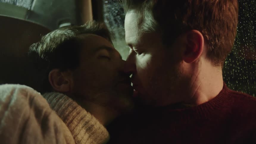 Men leaning against each other and hugging in the back seat turn and kiss as rain falls