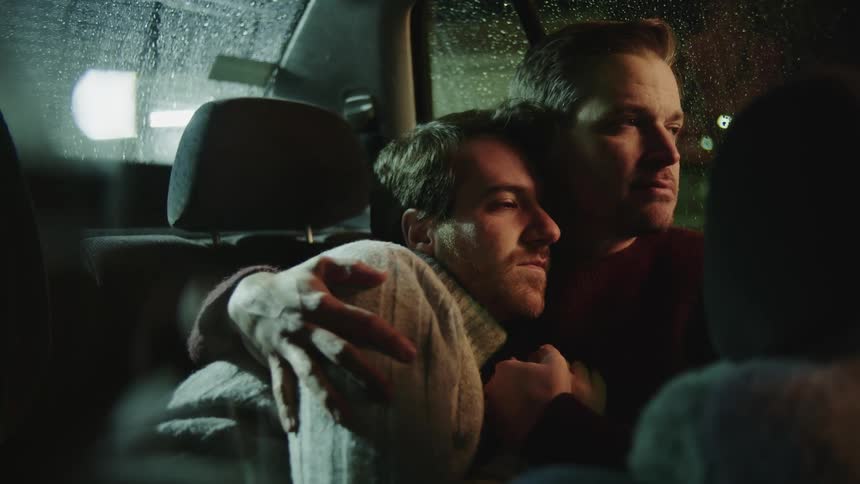 Two men sit in the back seat of a car and cuddle as one man rubs the others shoulder