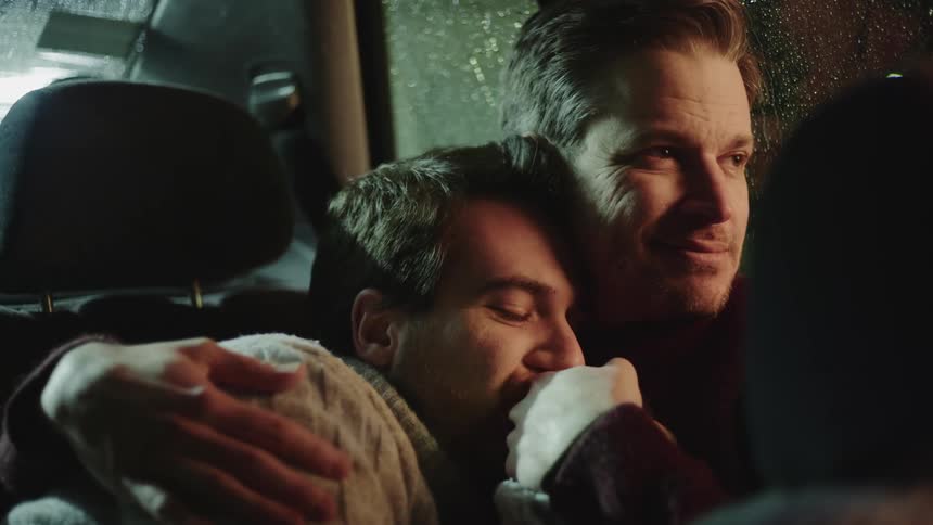 A man kisses the hand of another man while being embraced in the backseat of a vehicle on a rainy night