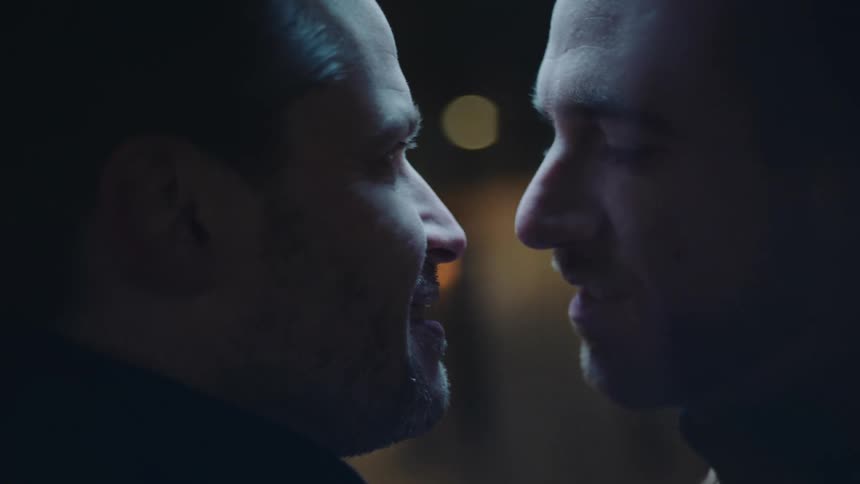 A man lovingly caresses and holds his mans face as they kiss and smile with city lights behind them