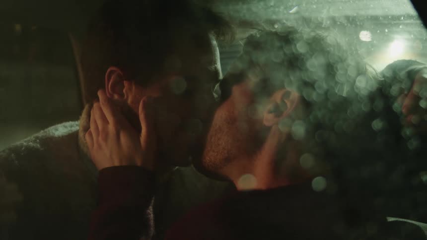 A pair of men kiss while sitting in the backseat of a vehicle on a rainy night