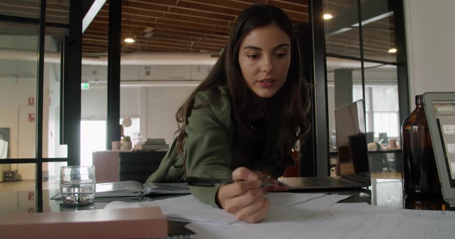 A young woman leans over a desk to write on a sheet of paper in an office during the day