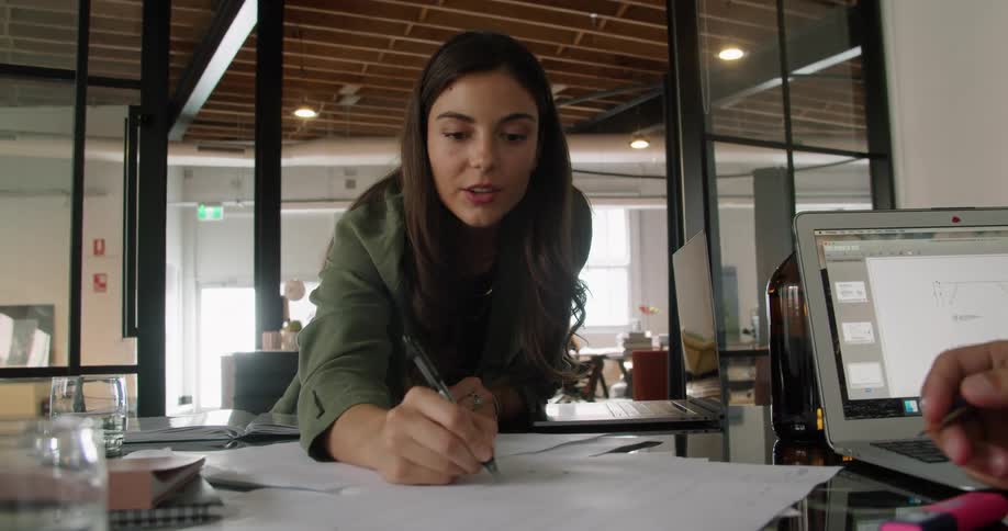 A woman leans cross her colleagues desk to write on a piece of paper as he sits and uses a laptop in his office