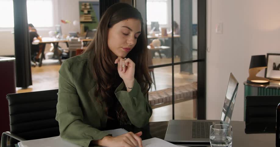 Woman sitting at the desk in the office reading sheets of data and typing on a laptop