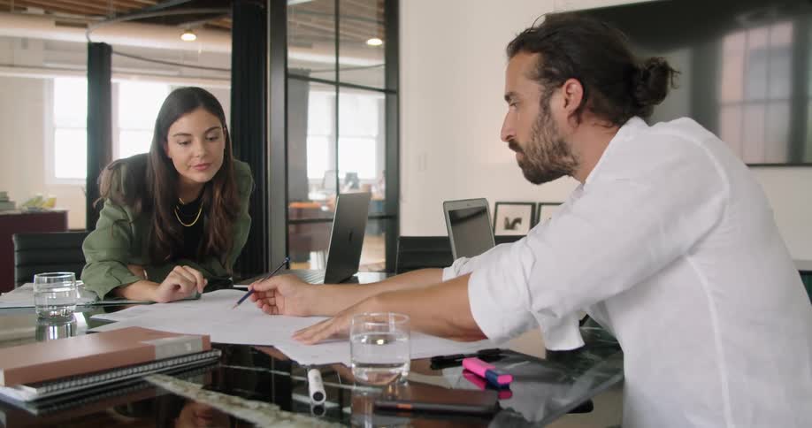 A man uses a pen to point lines on a paper out to a female coworker sitting across from him at a table in an office during the day