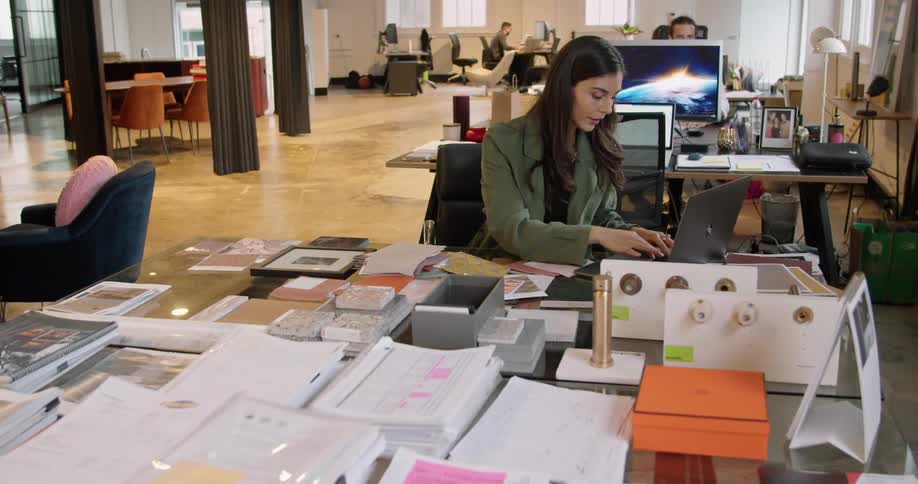 Woman working in an office sitting in a desk cluttered with samples typing on a laptop