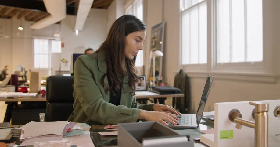 A woman sitting at a desk looking at paperwork and typing on a laptop while talking to herself