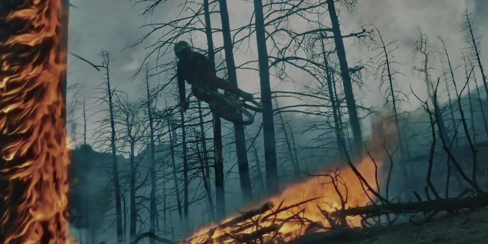 A person wearing a helmet rides a mountain bike over a pile of fallen trees in flames in a forest fire