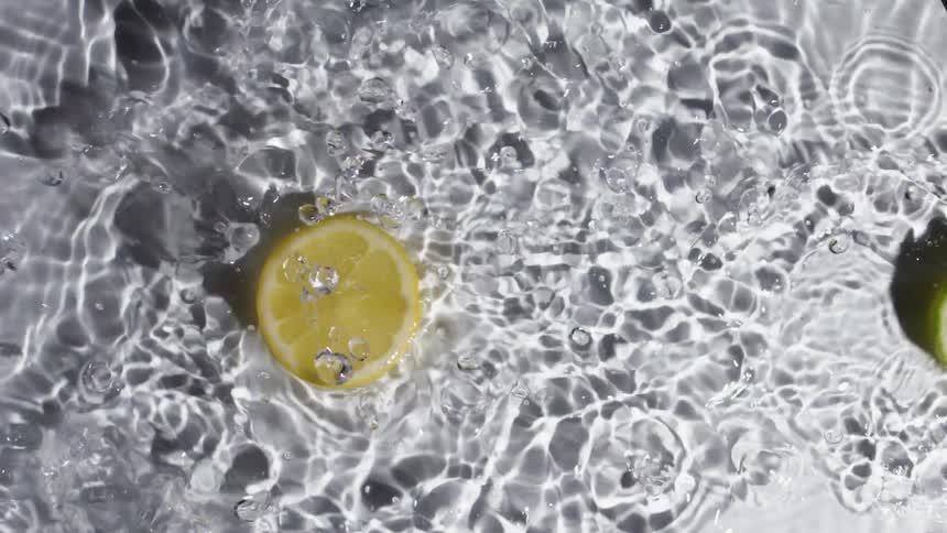 A slice of lemon and lime are tossed across the surface of clear water and cause splashing and ripples