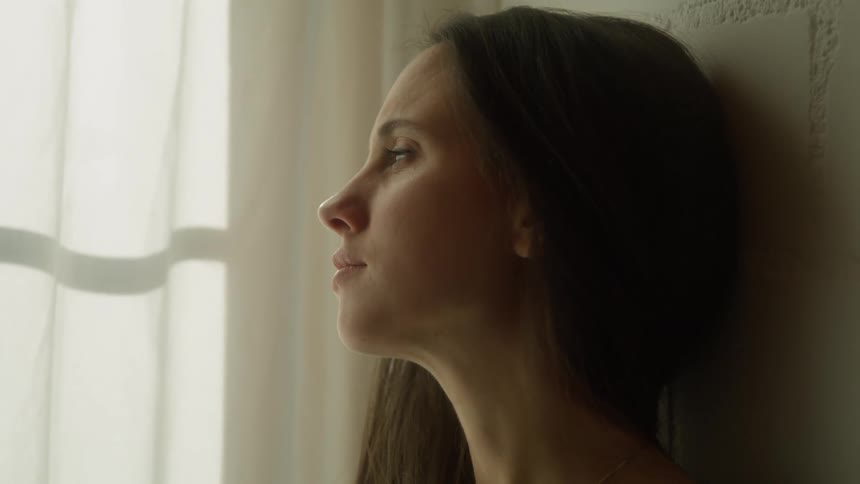 Woman with brown hair and a black shirt ponders thoughts next to the window in a room