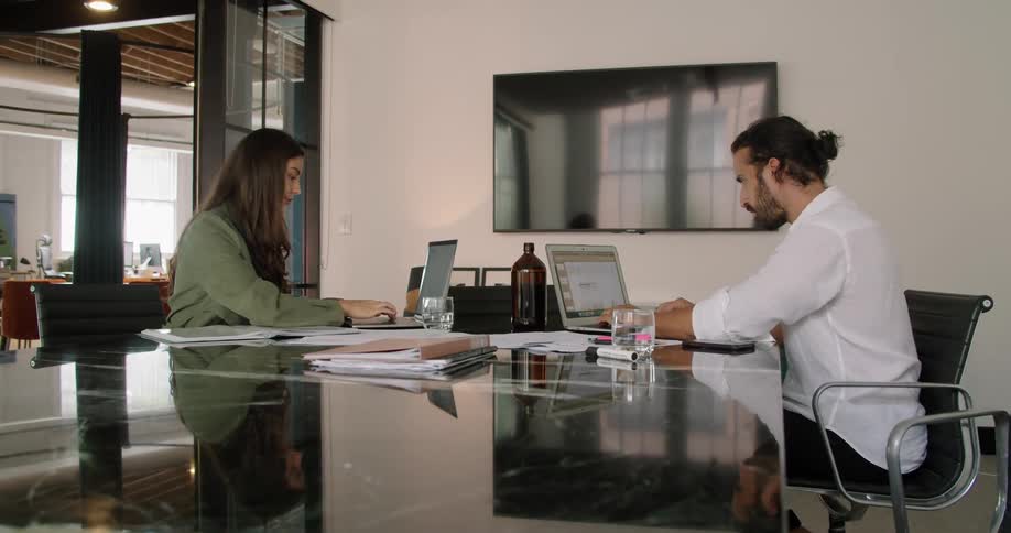 A man and a woman sitting on chairs at a table with laptops