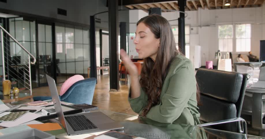A woman in an office sits at her desk and blows kisses and to someone on her laptop