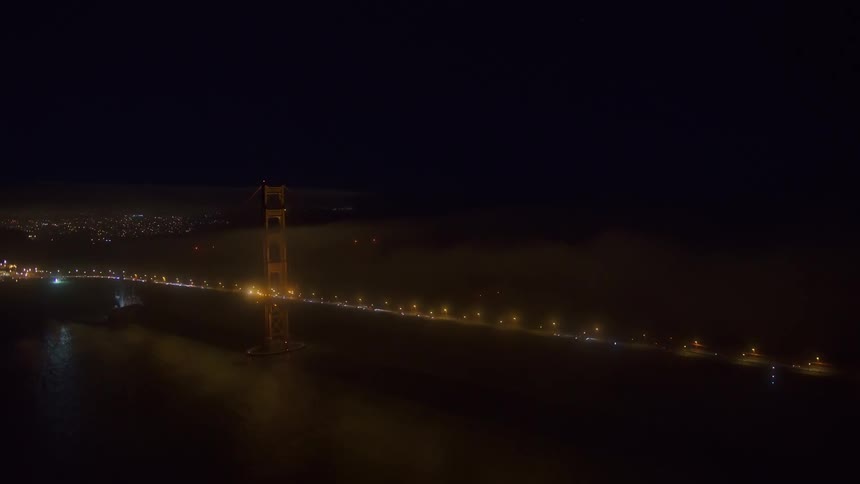 Traffic is heavy as dozens of vehicles cross a long bridge over a river near downtown at night