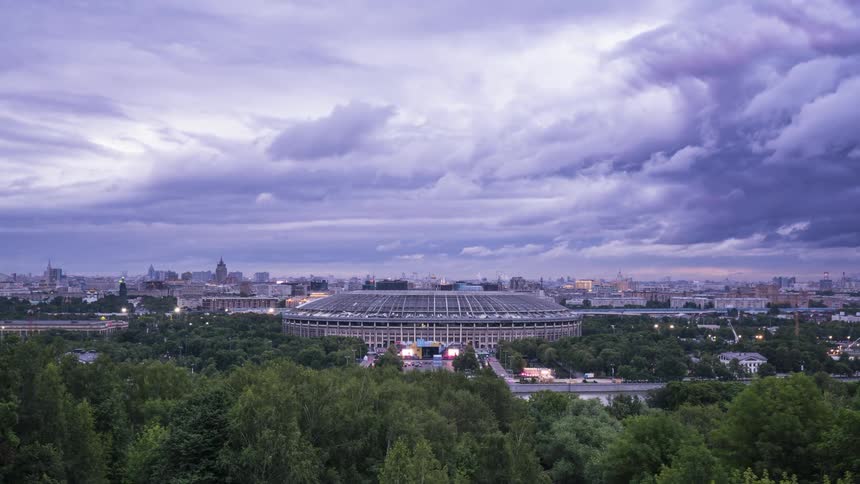 Lights glow from a stadium city buildings and traffic as clouds pass overhead